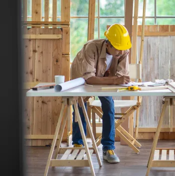 Top view of a professional architect in a yellow hard hat reviewing and sketching floor plans on blueprints with a pencil on a construction workbench, illustrating the detailed design and planning phase of Best Construction LA projects.